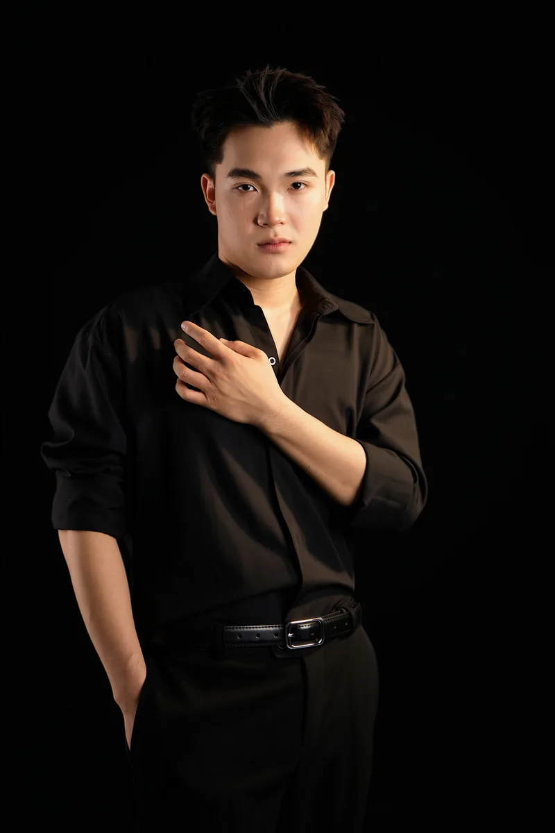 Ayden mặc sơ mi đen, một tay chạm ngực và một tay trong túi